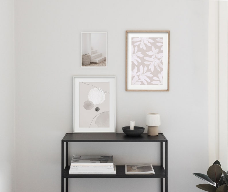 May include: Three framed prints on a white wall above a black metal shelf with books and a white ceramic candle holder. The prints are abstract art with circles and lines, a floral pattern, and a photograph of a white staircase.
