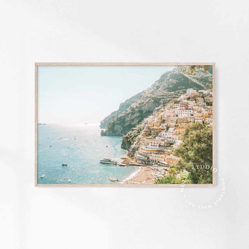 May include: A photograph of a colorful town built on a cliffside overlooking a blue ocean. The town is a mix of white, yellow, and orange buildings with green trees and plants. The ocean is a bright blue with whitecaps and a few boats.
