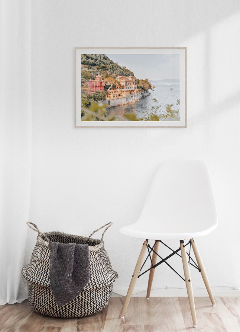 Pu&ograve; includere: Una fotografia incorniciata di un edificio colorato su una scogliera che si affaccia su un specchio d'acqua. L'edificio &egrave; di colore arancio chiaro e rosa con un tetto verde. L'acqua &egrave; di colore blu chiaro e in lontananza si vedono delle barche. La fotografia &egrave; incorniciata in una cornice di legno chiaro.