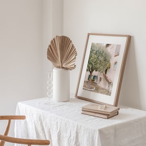 May include: A white table with a white linen tablecloth. A framed photograph of a tree in front of a building is on the table. A dried palm leaf is in a white vase on the table. Two books are stacked on top of the photograph.