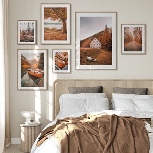 May include: Five framed autumn-themed prints hanging on a white wall above a bed with a brown blanket. The prints feature scenes of fall foliage, a lake with a boat, a cabin in the woods, and pumpkins.