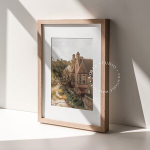 May include: A framed print of a vintage building with a red brick facade and a green roof. The building is surrounded by trees and foliage. The print is in a light wood frame.