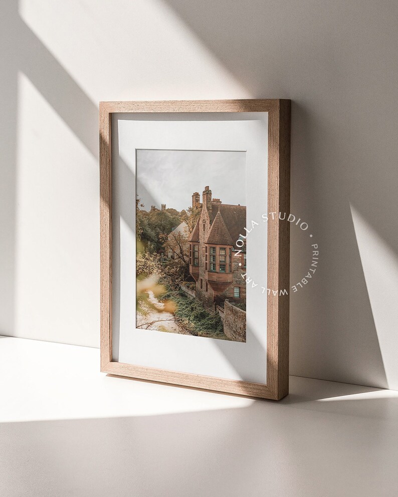May include: A framed print of a vintage building with a red brick facade and a green roof. The building is surrounded by trees and foliage. The print is in a light wood frame.