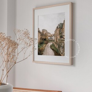 May include: A framed print of a photograph of a narrow river flowing through a village with stone buildings. The buildings are clustered together and have a rustic, medieval appearance. The river is surrounded by greenery and the sky is overcast.