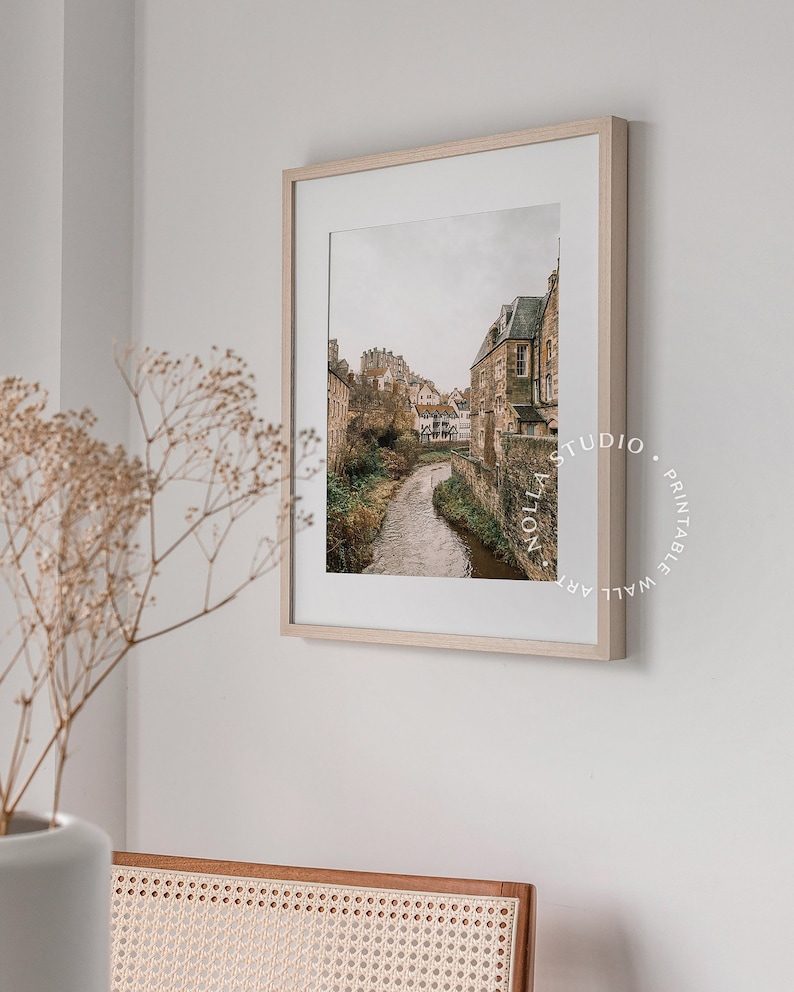 May include: A framed print of a photograph of a narrow river flowing through a village with stone buildings. The buildings are clustered together and have a rustic, medieval appearance. The river is surrounded by greenery and the sky is overcast.