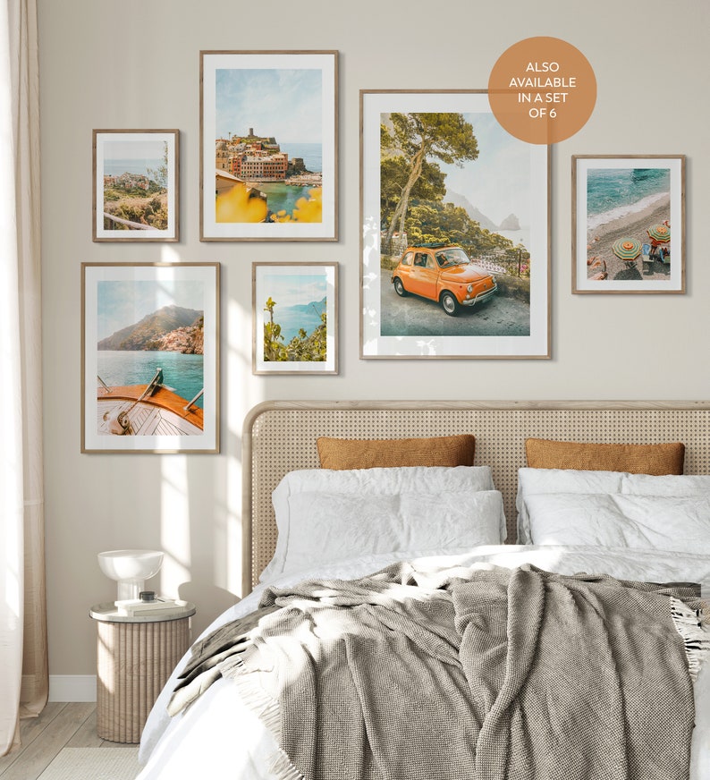 May include: A set of six framed prints featuring scenic views of the Italian coastline. The prints feature a variety of landscapes, including a colorful town, a winding road, a boat in the water, and a lush forest. The prints are all in a similar style, with a vintage, faded look. The prints are arranged in a grid on a wall above a bed with a white headboard and a gray blanket.