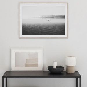 May include: A black and white photograph of a lake with a small boat in the distance. The lake is covered in fog and the sky is overcast. The photograph is framed in a light wood frame and is hanging on a white wall.