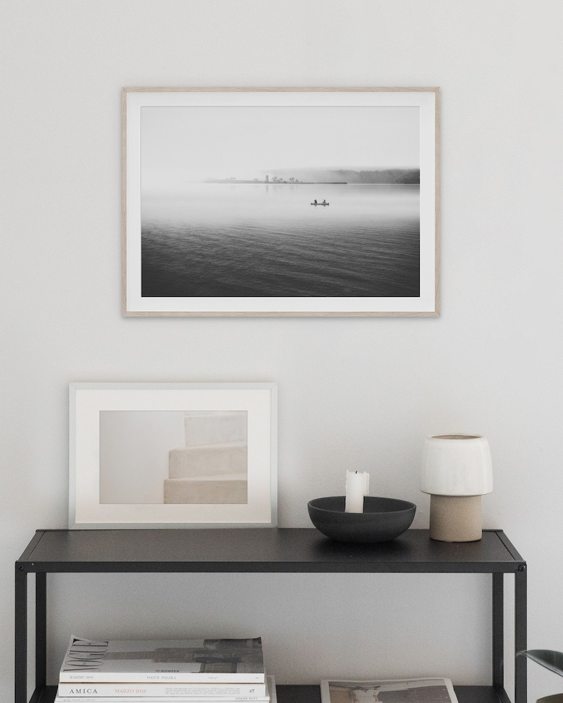 May include: A black and white photograph of a lake with a small boat in the distance. The lake is covered in fog and the sky is overcast. The photograph is framed in a light wood frame and is hanging on a white wall.