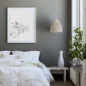 May include: A white bed with a white duvet cover and a white throw blanket. There is a white framed picture of a black and white floral print on the wall behind the bed. A white vase sits on a small wooden table next to the bed. A woven pendant light hangs from the ceiling.