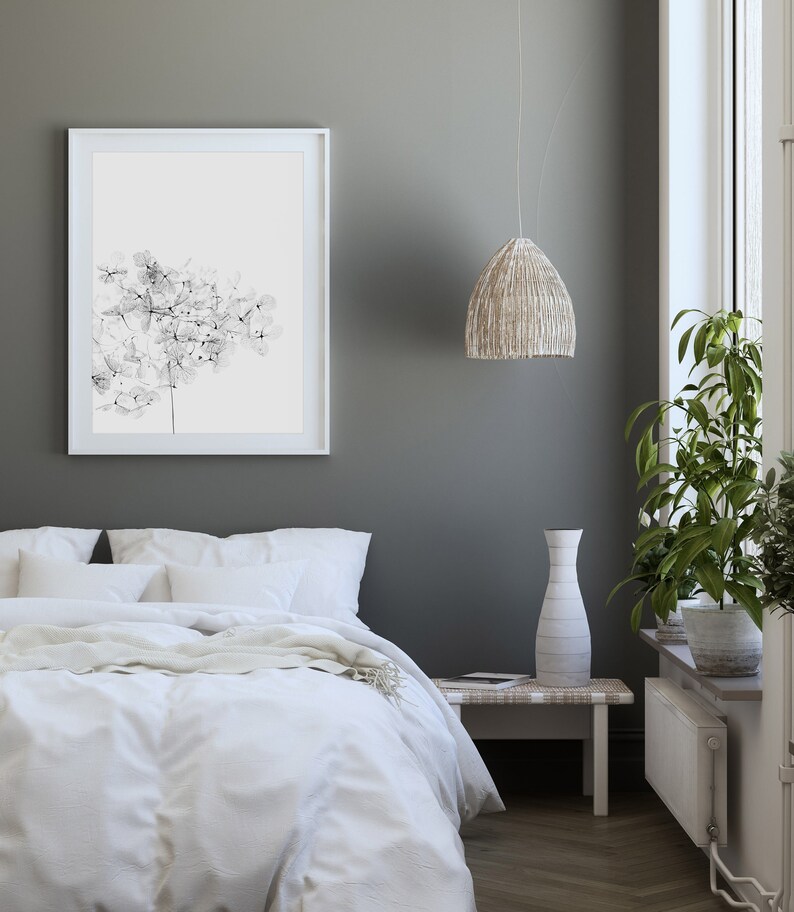 May include: A white bed with a white duvet cover and a white throw blanket. There is a white framed picture of a black and white floral print on the wall behind the bed. A white vase sits on a small wooden table next to the bed. A woven pendant light hangs from the ceiling.