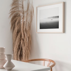 May include: A black and white photograph of a boat on a foggy lake with a cityscape in the background. The photograph is framed in a light wood frame and is hanging on a white wall. A tall vase with dried pampas grass is in the foreground.