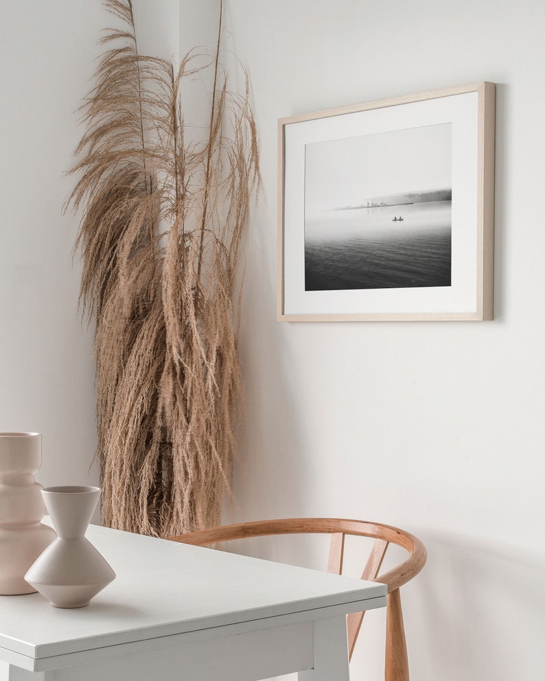 May include: A black and white photograph of a boat on a foggy lake with a cityscape in the background. The photograph is framed in a light wood frame and is hanging on a white wall. A tall vase with dried pampas grass is in the foreground.