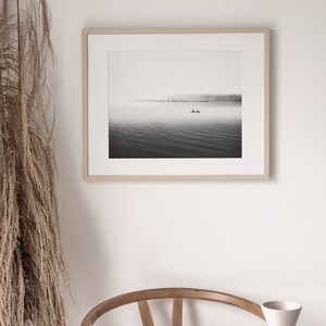May include: A black and white photograph of a lake with a small boat in the distance. The photo is framed in a light wood frame.