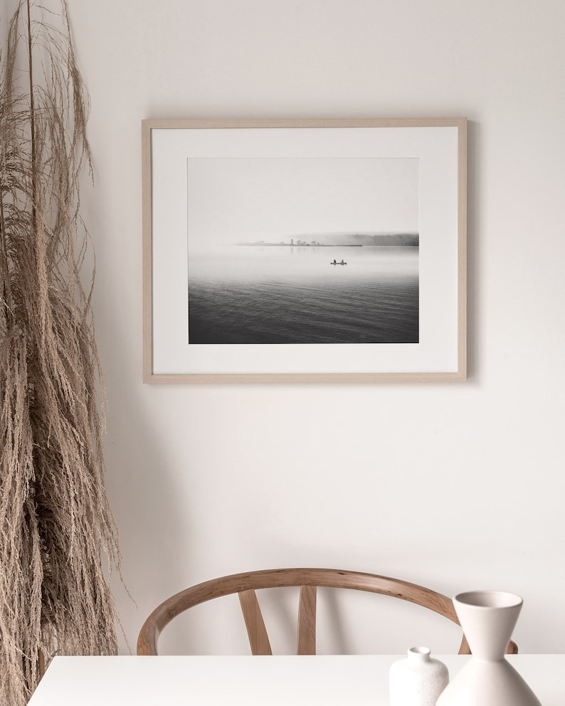 May include: A black and white photograph of a lake with a small boat in the distance. The photo is framed in a light wood frame.