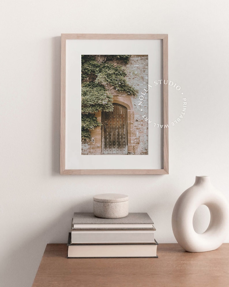 May include: A framed print of an old wooden door with ivy growing on the wall. The print is in a light wood frame and has a white mat. The print is titled "Nollas Studio. Printable Wall Art."