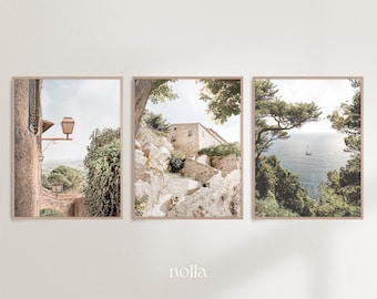 May include: A set of three framed prints featuring scenic views of a European village. The first print shows a stone building with a street lamp and a view of the surrounding countryside. The second print shows a stone staircase leading up to a building with a view of the surrounding countryside. The third print shows a view of the ocean from a cliff with a sailboat in the distance.