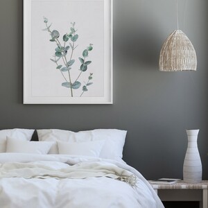 May include: A white bed with white bedding and a white throw blanket. A framed print of a green eucalyptus branch hangs on the wall above the bed. A white vase and a wicker lamp hang above a white nightstand.