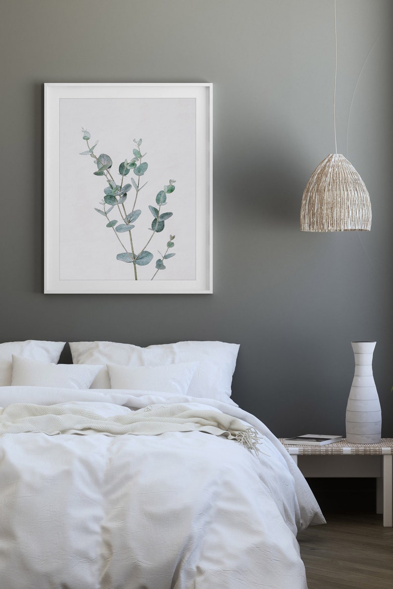 May include: A white bed with white bedding and a white throw blanket. A framed print of a green eucalyptus branch hangs on the wall above the bed. A white vase and a wicker lamp hang above a white nightstand.