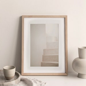 Peut inclure: Une photographie encadr&eacute;e d'un escalier blanc avec un cadre en bois clair. Le cadre est pos&eacute; sur une table blanche avec un vase en c&eacute;ramique blanche et une petite tasse en c&eacute;ramique.