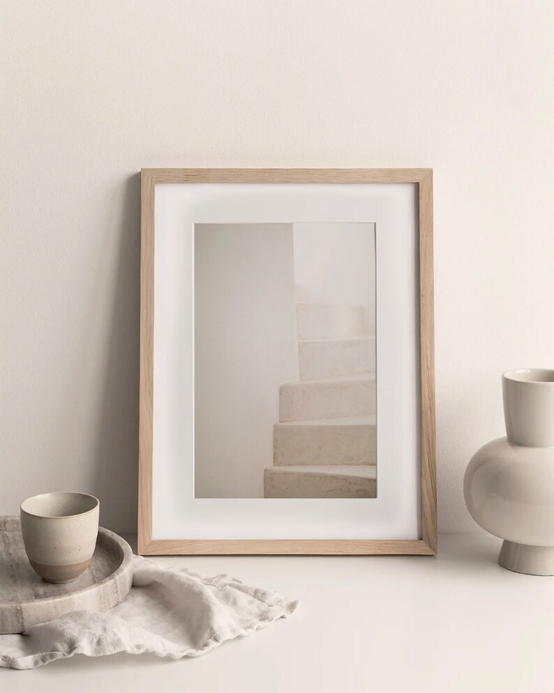 Peut inclure: Une photographie encadr&eacute;e d'un escalier blanc avec un cadre en bois clair. Le cadre est pos&eacute; sur une table blanche avec un vase en c&eacute;ramique blanche et une petite tasse en c&eacute;ramique.