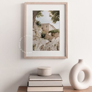 May include: A framed photograph of a stone building with a staircase leading up to it. The building is surrounded by trees and greenery. The photograph is in a light wood frame and is hanging on a white wall.