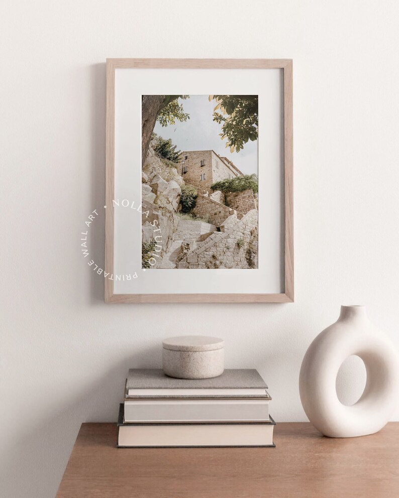 May include: A framed photograph of a stone building with a staircase leading up to it. The building is surrounded by trees and greenery. The photograph is in a light wood frame and is hanging on a white wall.