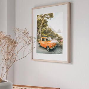 May include: A framed photograph of an orange vintage car parked on a winding road with a view of the ocean and a lush green forest in the background.