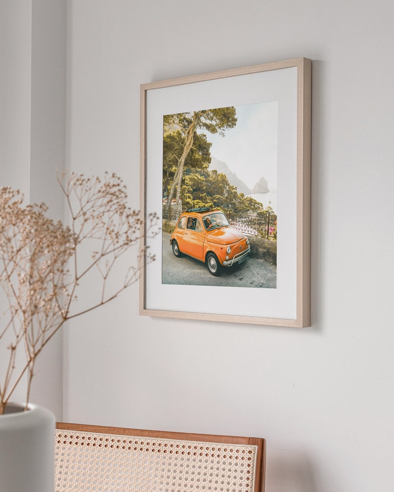 May include: A framed photograph of an orange vintage car parked on a winding road with a view of the ocean and a lush green forest in the background.