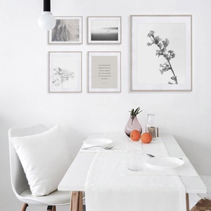 May include: A white dining table with a white tablecloth and runner. There are two white plates, a glass, and two oranges on the table. There are four framed black and white prints on the wall behind the table. The prints feature a landscape, a floral design, a tree branch, and a quote.