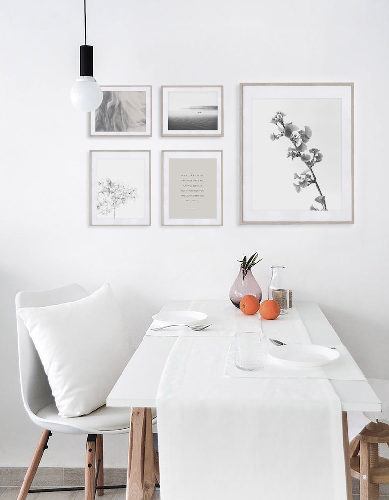 May include: A white dining table with a white tablecloth and runner. There are two white plates, a glass, and two oranges on the table. There are four framed black and white prints on the wall behind the table. The prints feature a landscape, a floral design, a tree branch, and a quote.
