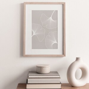 Puede incluir: Una impresi&oacute;n enmarcada con tres contornos de hojas de ginkgo blancas sobre un fondo gris claro. La obra de arte est&aacute; en un marco de madera clara y se exhibe encima de una pila de libros, un peque&ntilde;o recipiente redondo y un jarr&oacute;n blanco con una abertura circular.