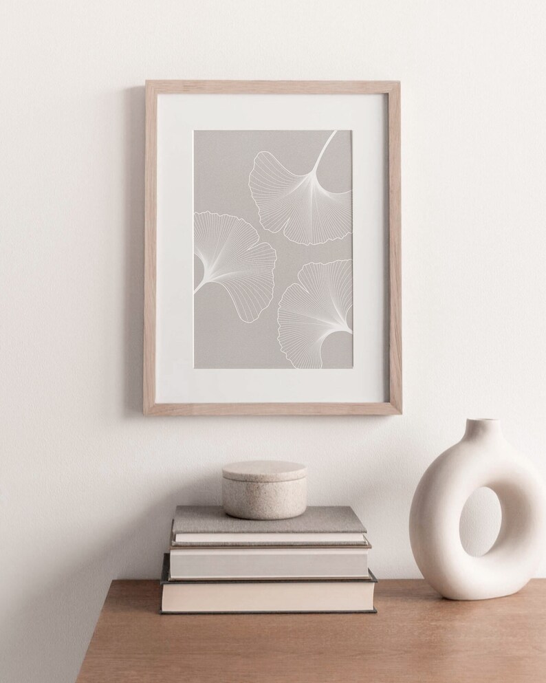 Puede incluir: Una impresi&oacute;n enmarcada con tres contornos de hojas de ginkgo blancas sobre un fondo gris claro. La obra de arte est&aacute; en un marco de madera clara y se exhibe encima de una pila de libros, un peque&ntilde;o recipiente redondo y un jarr&oacute;n blanco con una abertura circular.