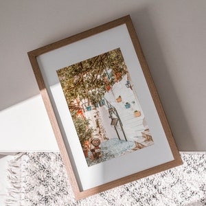 May include: A framed photograph of a narrow, cobblestone street in a European city. The street is lined with white buildings and lush greenery. A small, red scooter is parked on the street.