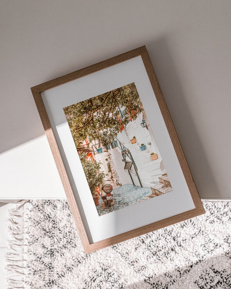 May include: A framed photograph of a narrow, cobblestone street in a European city. The street is lined with white buildings and lush greenery. A small, red scooter is parked on the street.