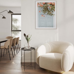 May include: A white, fluffy armchair with a gold base sits in a living room with a side table and a framed print of a white door with pink flowers hanging over it.