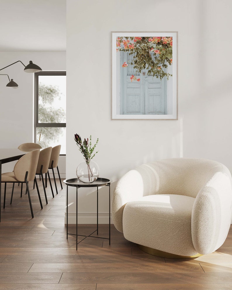May include: A white, fluffy armchair with a gold base sits in a living room with a side table and a framed print of a white door with pink flowers hanging over it.