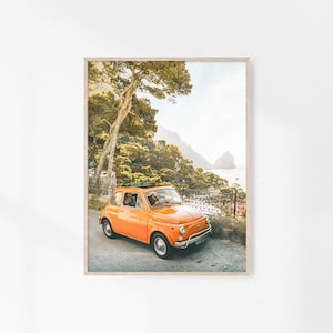 May include: A bright orange vintage Fiat car parked on a winding road with a view of a rocky coastline and lush green trees.