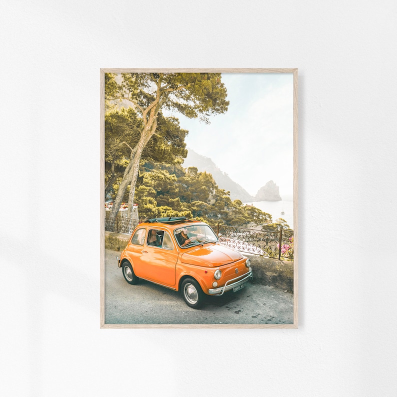 May include: A bright orange vintage Fiat car parked on a winding road with a view of a rocky coastline and lush green trees.
