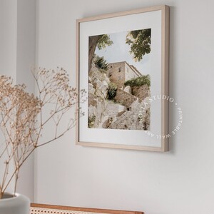 May include: A framed photograph of a stone building with a staircase leading up to it. The building is surrounded by trees and greenery. The photograph is in a light wood frame.