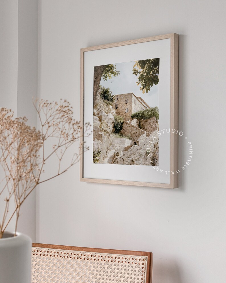 May include: A framed photograph of a stone building with a staircase leading up to it. The building is surrounded by trees and greenery. The photograph is in a light wood frame.