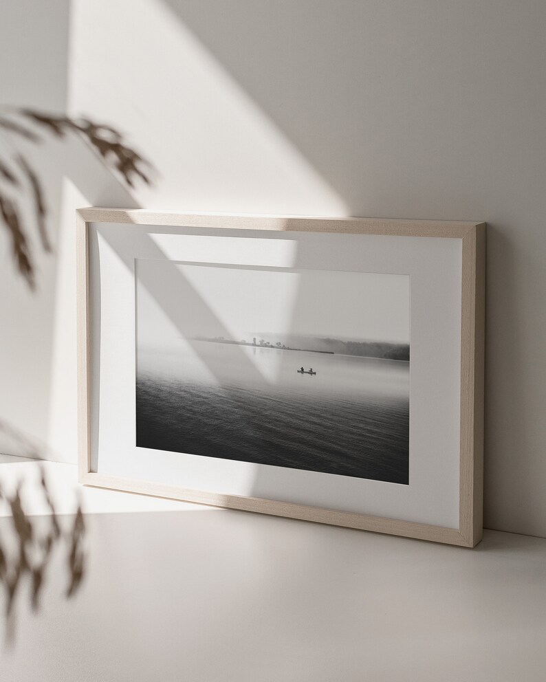May include: A black and white photograph of a lake with a small boat in the distance. The photo is framed in a light wood frame.