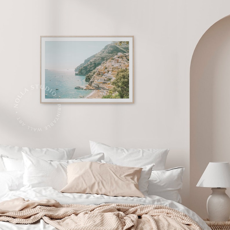May include: A framed photograph of a coastal town built on a hillside overlooking the ocean. The town is a vibrant mix of colorful buildings and lush greenery. The water is a clear blue, and the sky is a soft, hazy blue.