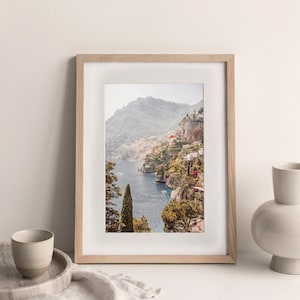 May include: A framed photograph of a coastal town nestled into a hillside overlooking a blue body of water. The town is built on a steep cliff with lush green trees and foliage. The photo is in a light wood frame.