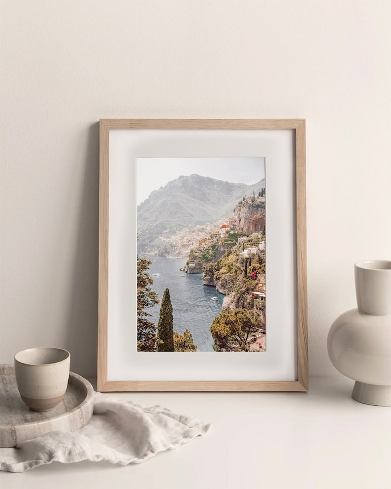 May include: A framed photograph of a coastal town nestled into a hillside overlooking a blue body of water. The town is built on a steep cliff with lush green trees and foliage. The photo is in a light wood frame.