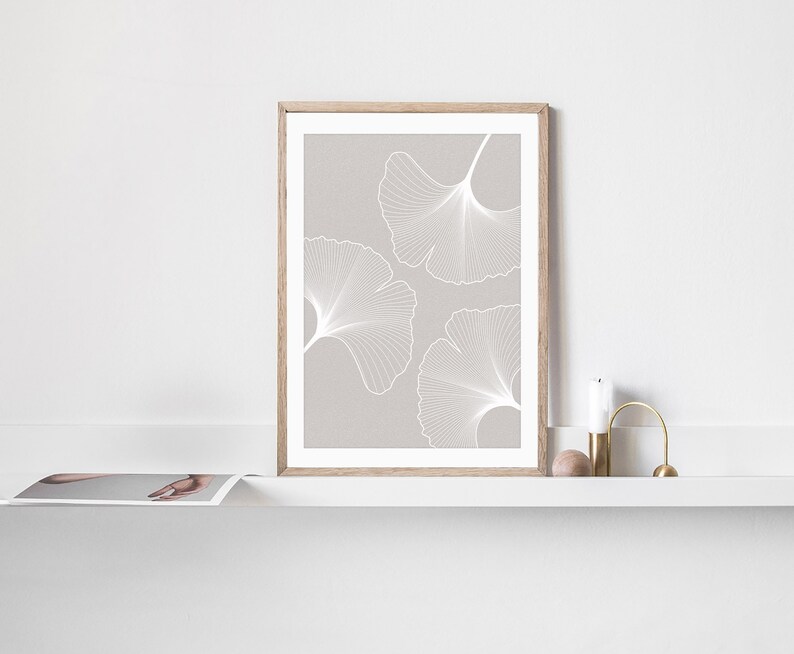 Puede incluir: Una impresi&oacute;n enmarcada con tres dise&ntilde;os de hojas de ginkgo blancas sobre un fondo gris claro. La obra de arte se muestra en un marco de madera clara. La impresi&oacute;n se exhibe en una estanter&iacute;a blanca con una vela y objetos decorativos.