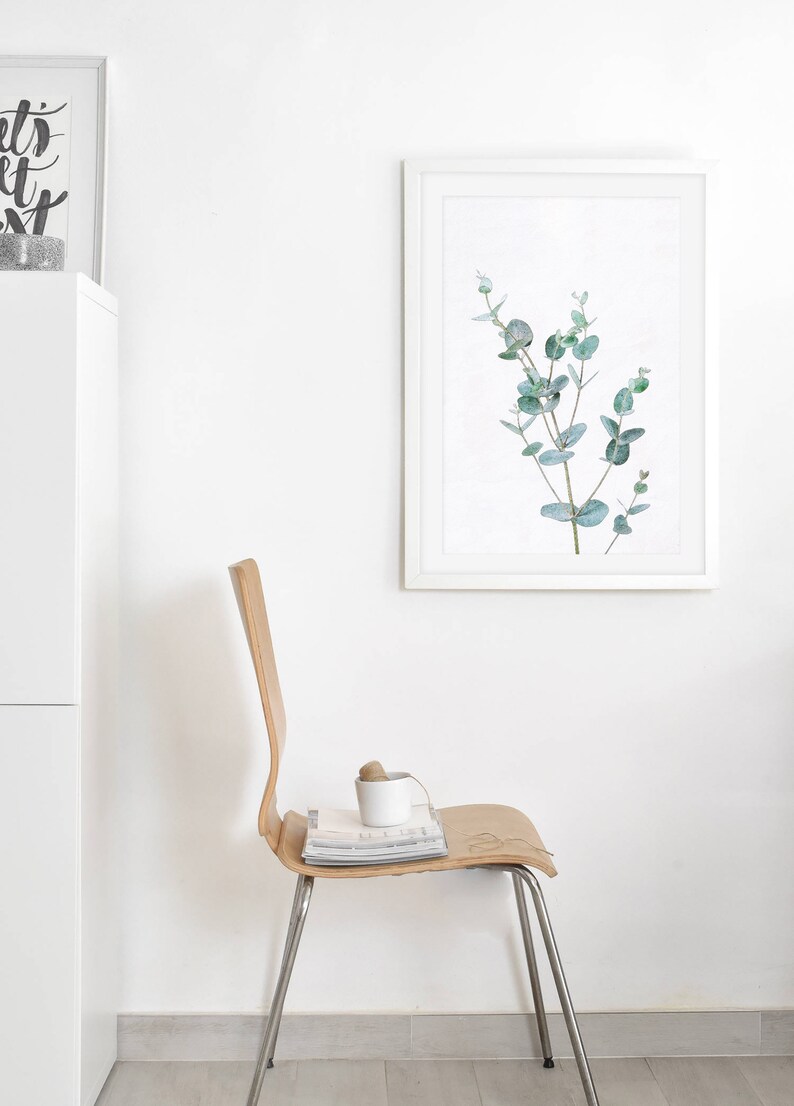 May include: A white framed print of a watercolor painting of a eucalyptus branch with green leaves against a white background. A wooden chair with a magazine and a white cup on the seat. The chair is in front of a white wall.