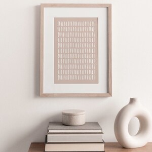 May include: A framed print with a beige background and white abstract lines. The print is hanging on a white wall above a stack of books and a white ceramic vase with a ring-shaped opening.