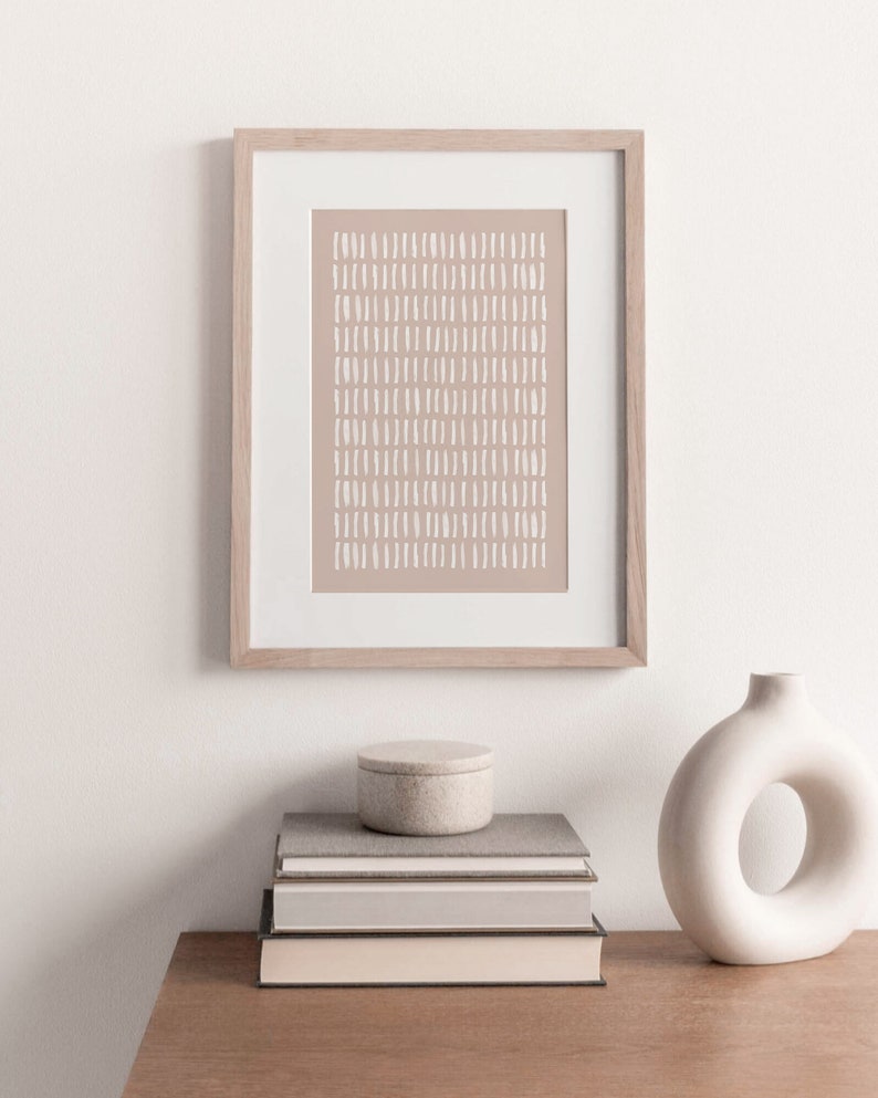 May include: A framed print with a beige background and white abstract lines. The print is hanging on a white wall above a stack of books and a white ceramic vase with a ring-shaped opening.