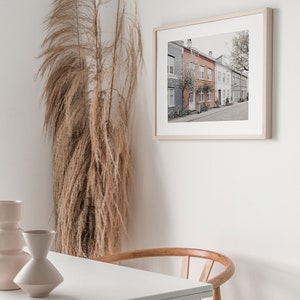 May include: A framed print of a row of colorful houses with a tree in the foreground. The houses are painted in shades of pink, blue, and gray. The print is hanging on a white wall.