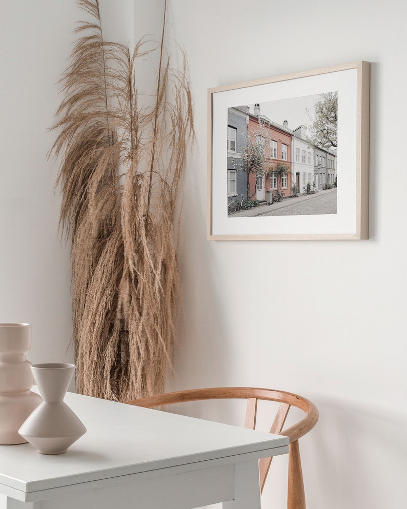 May include: A framed print of a row of colorful houses with a tree in the foreground. The houses are painted in shades of pink, blue, and gray. The print is hanging on a white wall.